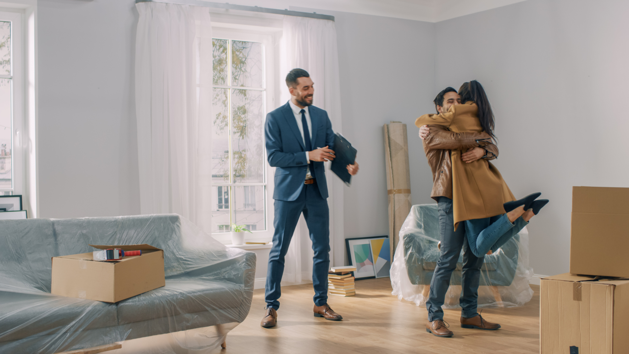 A real-estate agent smiles as a couple embraces joyfully in their new home surrounded by moving boxes.